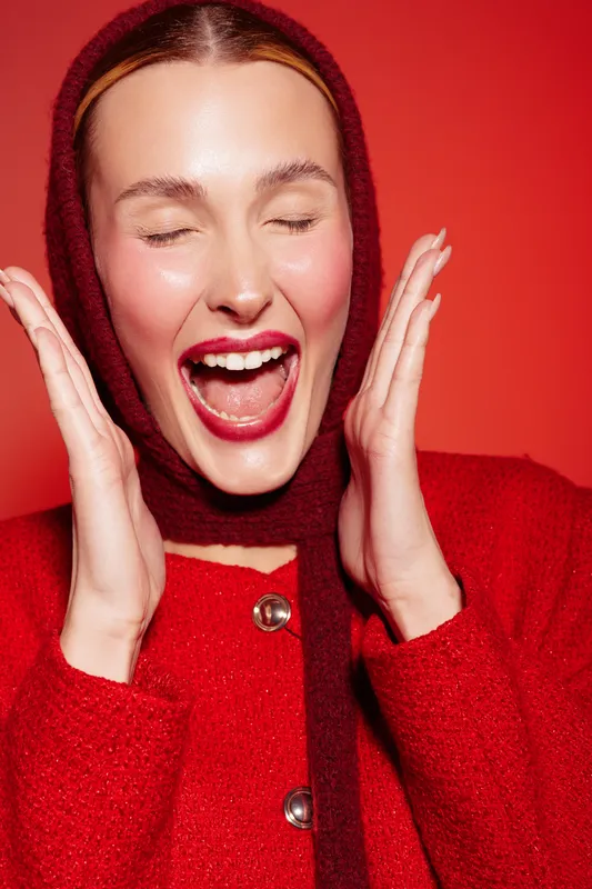 Happy woman in red sweater and scarf laughs with eyes closed on red background. Concept of joy, fashion, winter clothing, and positive emotions in studio portrait.