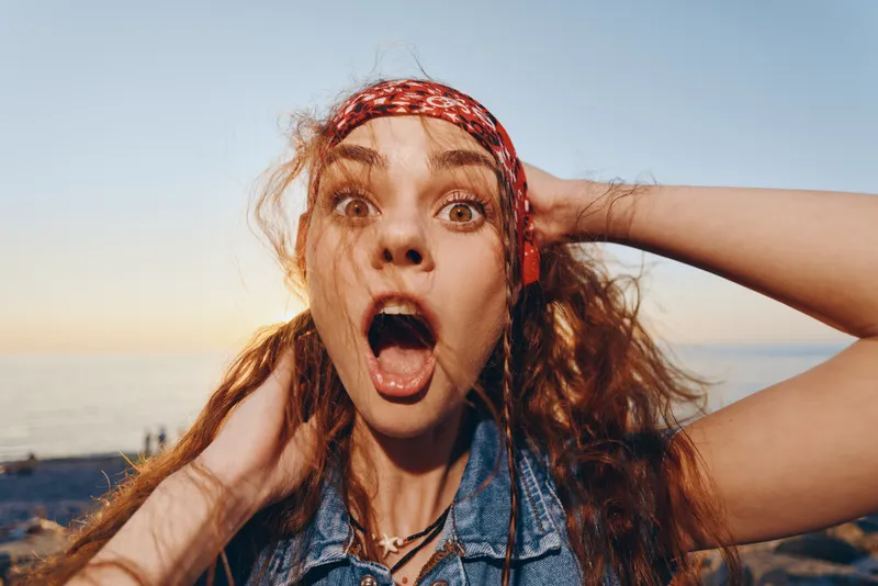 surprised woman in modern boho-western denim street style with expressive face and film color effect outdoors near sunset sky