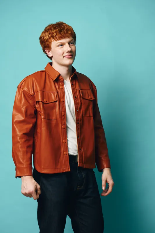 Young man with red hair wearing brown leather jacket and white shirt standing against blue background looking thoughtful and confident in casual fashion style for portrait.