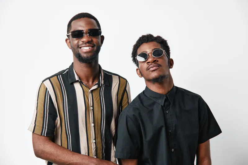 Two young black men with dark skin wearing sunglasses and casual shirts posing together smiling and confident on white background studio portrait.