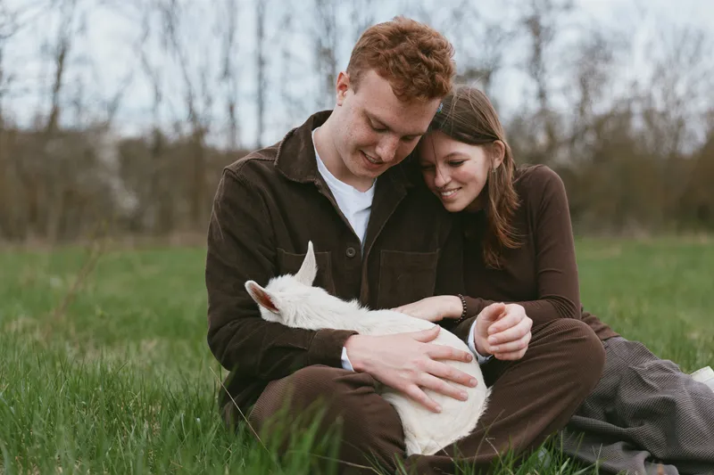 Loving couple sitting on a grassy field, gently holding a goat kid, enjoying outdoor life. Cozy, intimate moment capturing affection, nature, and simple happiness in a relaxed lifestyle scene.