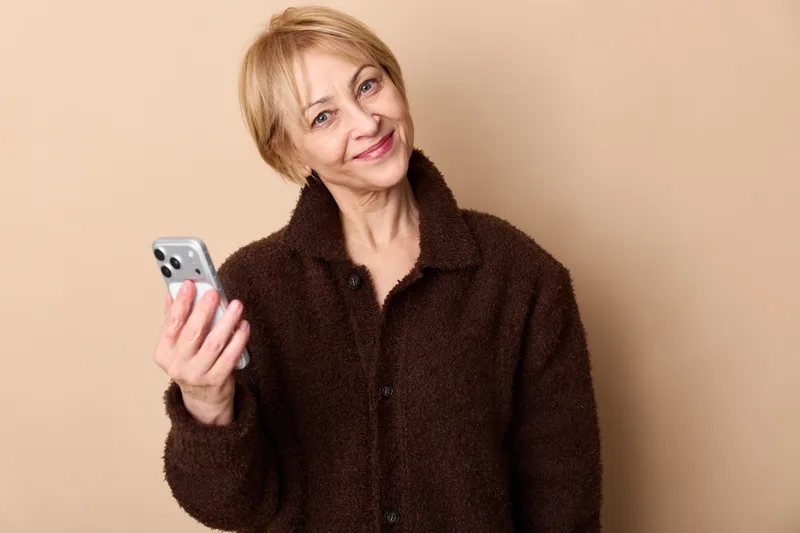 Woman with smartphone, smiling warmly in studio portrait, elderly, short blonde hair, wearing brown jacket, cozy fleece texture, indoor beige background, relaxed pose