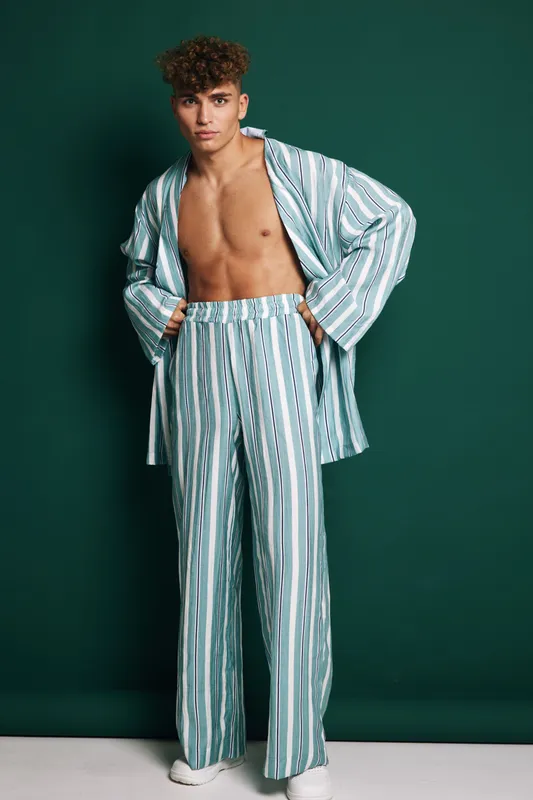 Young man with curly hair wearing striped loose pants and open shirt posing barefoot against dark green background in studio for fashion and casual outfit style photography.