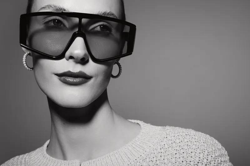 Young white woman with pale skin wears large black sunglasses and hoop earrings. She poses with a slight smile against a plain background in monochrome style.