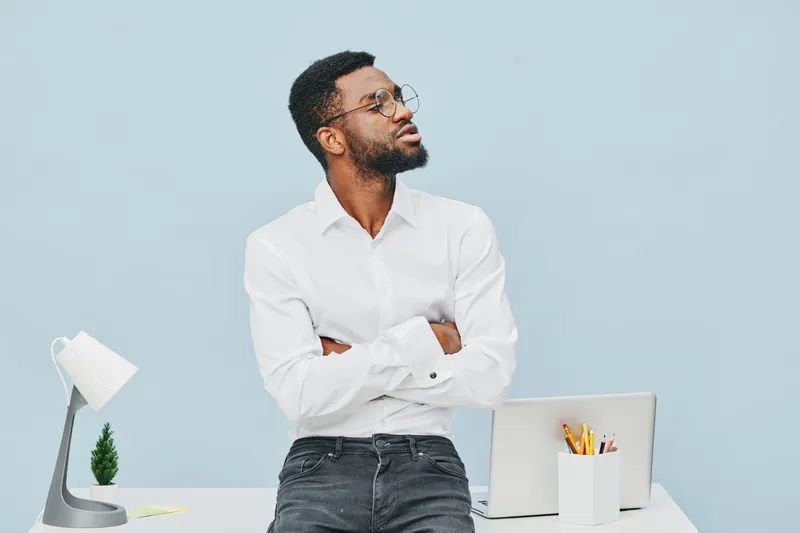 Thoughtful young man with glasses in white shirt standing with crossed arms at modern office desk on light blue background. Workplace professional confident casual style. People lifestyle concept