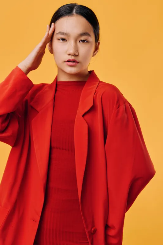 Young woman with long dark hair wearing bright red jacket and sweater posing with hand near face on yellow background studio portrait. Fashion, beauty, model, casual and confident style concept.
