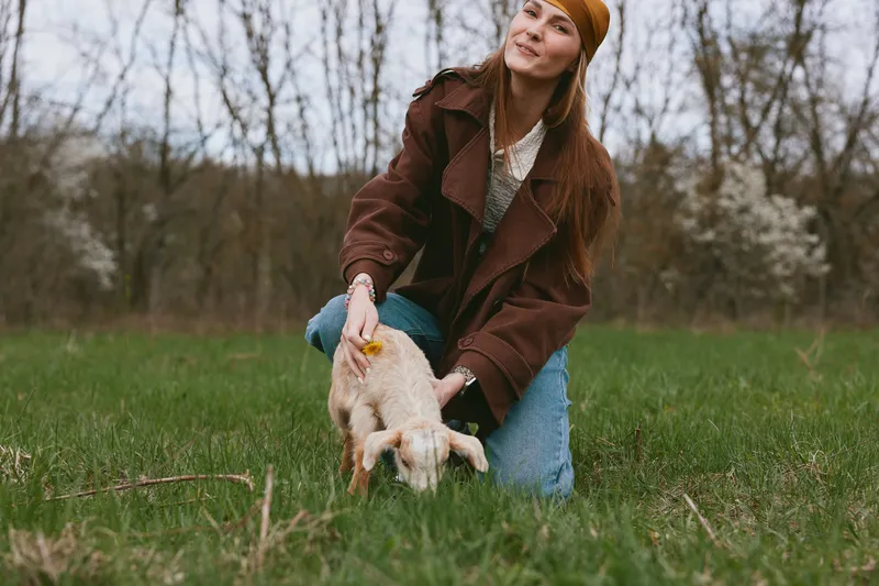 Friendly woman in a brown coat kneels on green grass, guiding a playful goat kid. Outdoor lifestyle scene with natural light, showing care, companionship, and a rustic countryside mood.