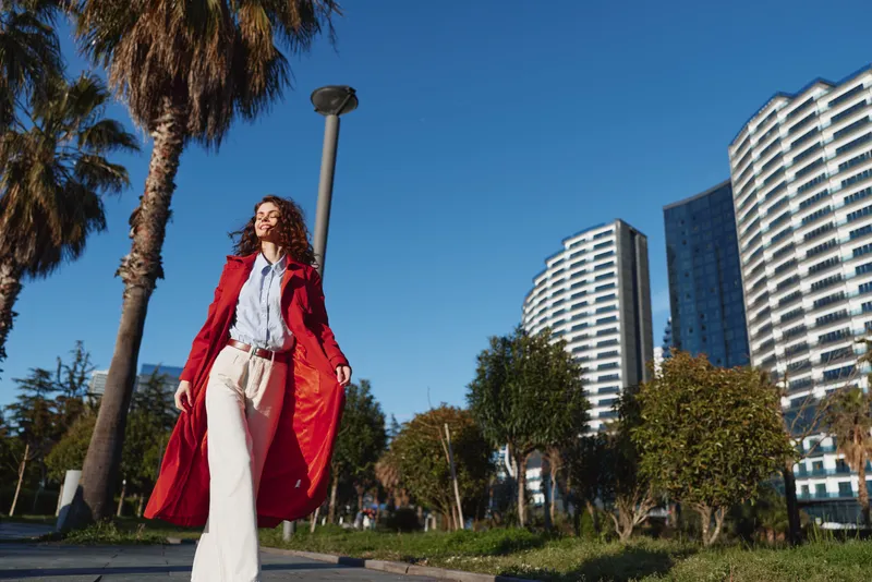Young woman with curly hair walks confidently along a sunny urban street wearing a long red coat and white pants under a clear blue sky.