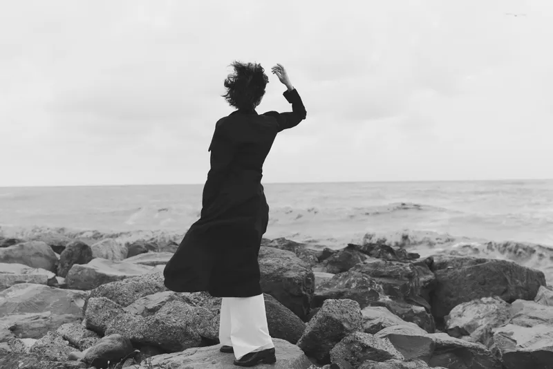 Woman on rocks by the sea gazing toward horizon, wind in coat, monochrome scene capturing resilience, exploration, and solitary contemplation on rugged shoreline
