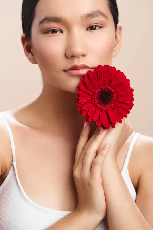Asian woman gazes softly at the camera as she holds a bright red flower near her face, conveying calm elegance and beauty against a neutral backdrop.