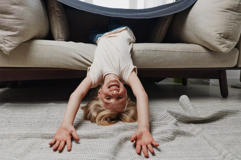 Happy child hanging upside down from couch with arms stretched on carpet. Playful kid in casual clothes smiling indoors. Concept of childhood joy, fun, and home leisure activity.