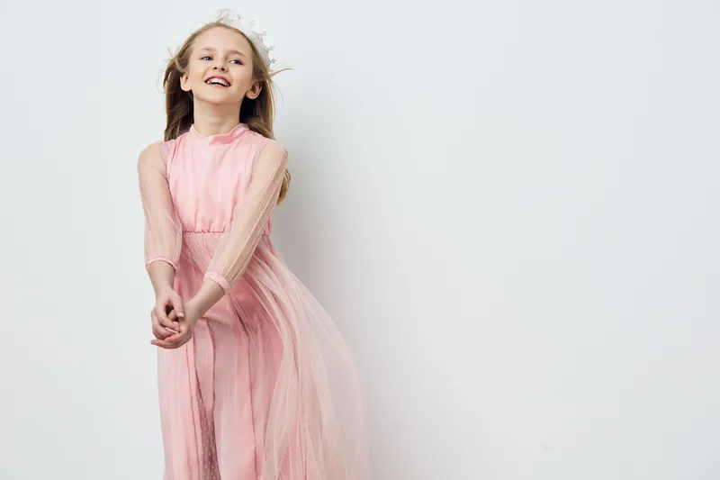 Cheerful young girl in a flowing pink dress smiles joyfully against a minimalist light background, embodying childhood innocence and playfulness