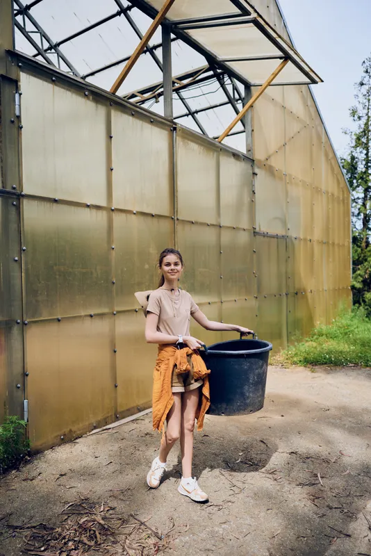 Woman girl greenhouse bucket gardening gardener outdoor farm scene carrying black pail by translucent panels, casual summer outfit with tied sweatshirt and sneakers in rural horticulture