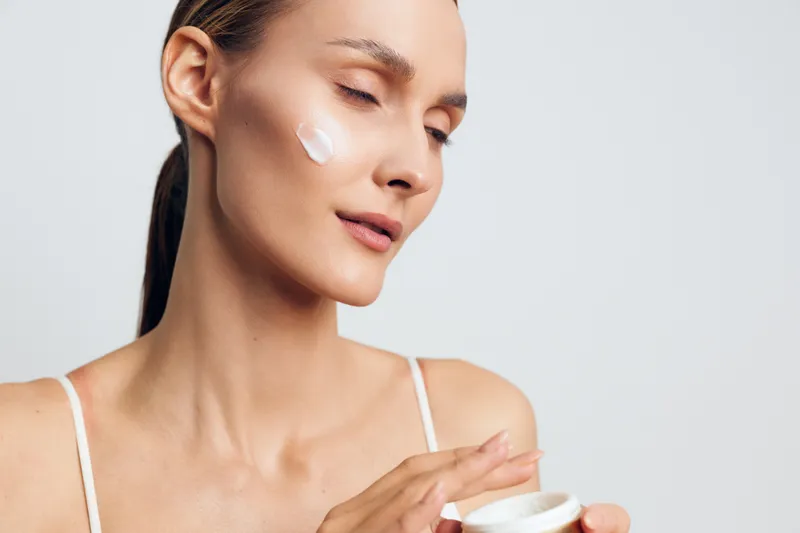 Woman skincare beauty close up applying cream on face with cotton pad, calm gaze, soft studio light, minimal makeup, clean gray background, moisturizing skin care, daily hydration and glow