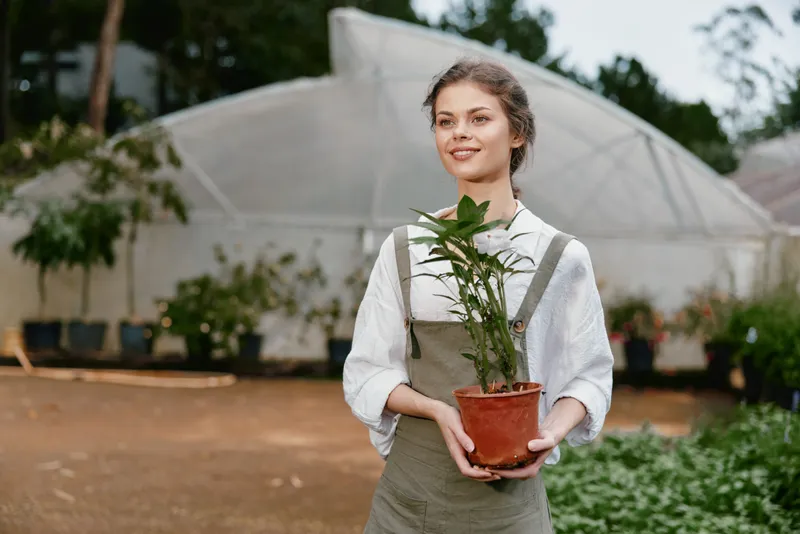Young woman in green overalls smiling while holding a potted plant in a greenhouse, symbolizing gardening, nature, and sustainable living
