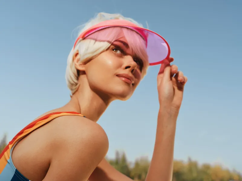A stylish young woman wearing a pink visor gazes toward the distance under a bright blue sky, capturing a sunny, athletic moment outdoors.