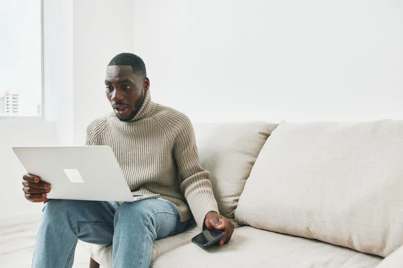 Young Black man sitting on a sofa, using a laptop and smartphone, dressed in a cozy sweater, looking surprised or excited in a modern interior
