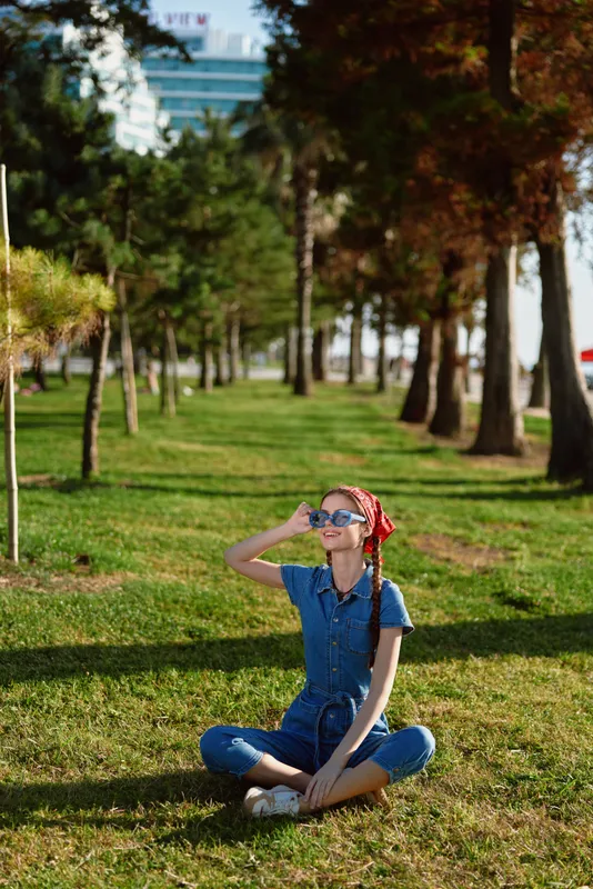 Lifestyle woman in modern boho-western denim street style sits cross-legged on green grass wearing sunglasses and a red headscarf under warm natural sunlight, film color.