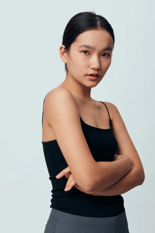 Young woman with clear skin and black hair wearing black tank top standing with crossed arms against light background, looking confident and natural in minimalistic style.