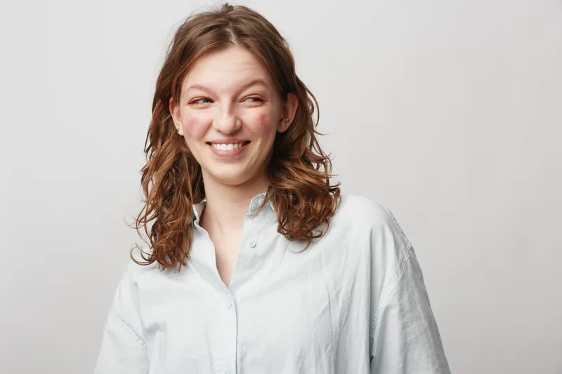 Smiling young woman with curly brown hair wearing white shirt looks to side with playful expression. Studio portrait on light background. Concept of happiness, youth, and casual style.