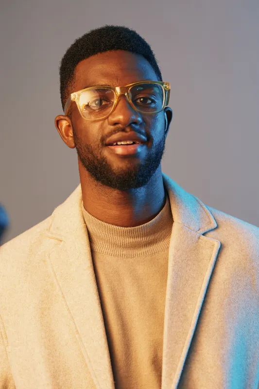 Stylish young man in glasses wearing a cozy sweater and a light coat, exuding confidence with a warm smile against a soft gradient background