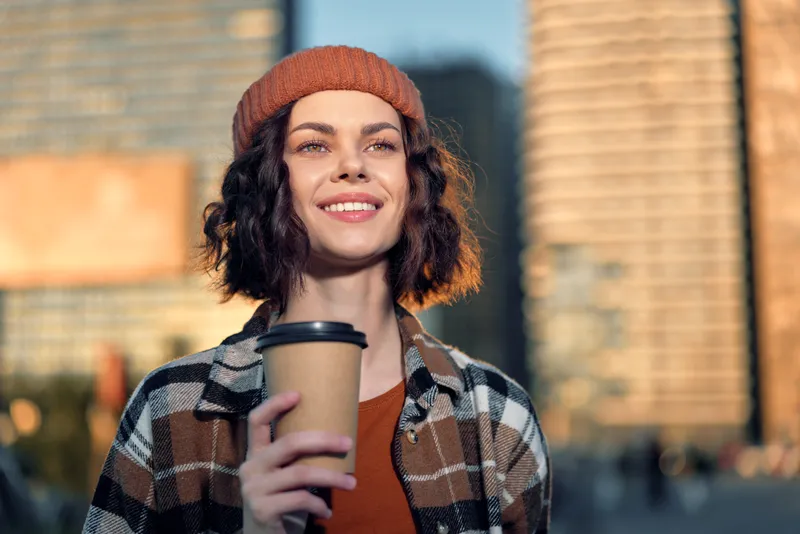 woman coffee smile urban beanie sunlight portrait candid Young woman holding takeaway cup on city street at golden hour glow, authenticity and mindful living captured in emotional storytelling mood.