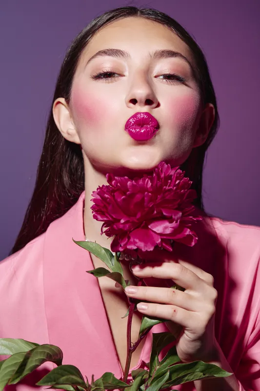 Young woman with vivid fuchsia lipstick and heavy pink blush poses with dark pink flower on purple background in pink fashionable attire beauty creative concept.