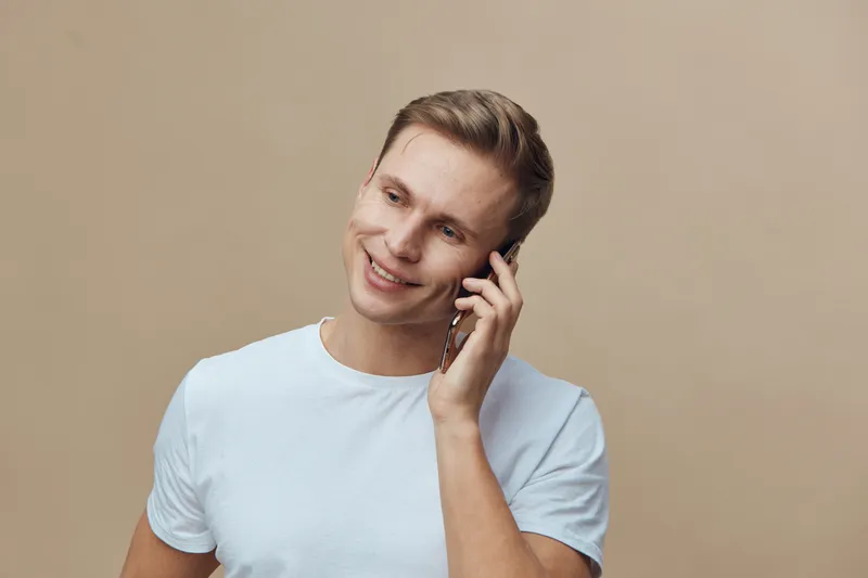Smiling young man with light brown hair wearing white t-shirt talking on mobile phone, looking away, cheerful expression, casual style, isolated on beige background, studio portrait, lifestyle concept