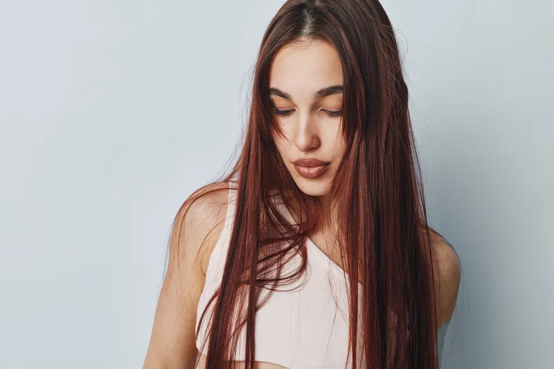 Young woman with long straight hair and natural makeup looking downwearing a white sleeveless top against plain light background with calm and thoughtful expression.