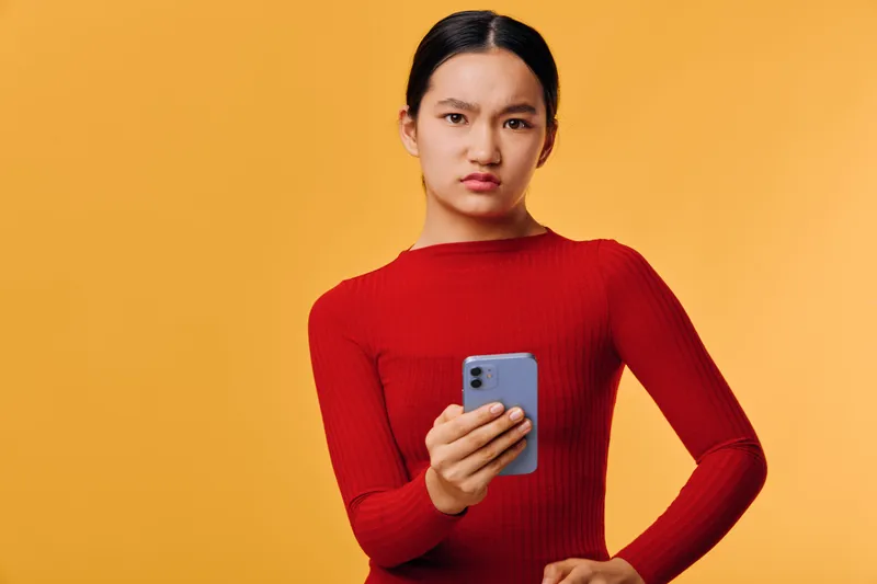 Young woman with serious face holding smartphone looking at camera in red sweater on yellow background. Mobile device, technology and communication concept with confident expression.