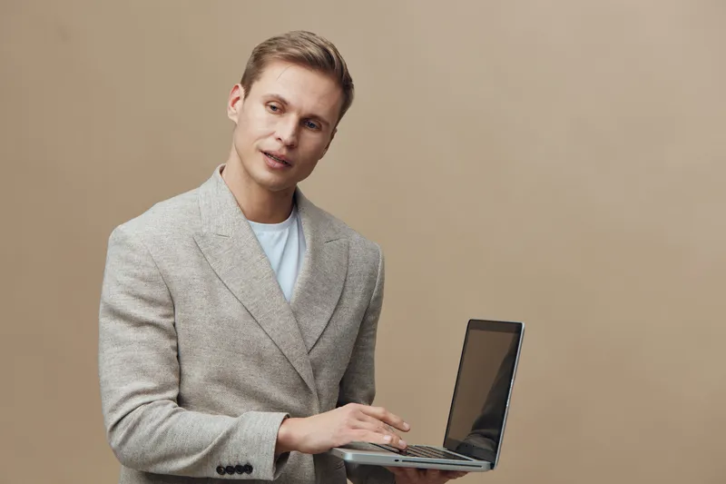 young man holding laptop standing in beige jacket looking aside digital nomad technology concept in studio portrait