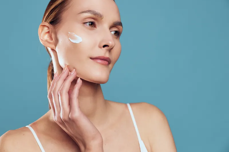 Young woman applies moisturizing cream on her face with fingers, showing skincare routine and healthy skin care on blue background in close up portrait.
