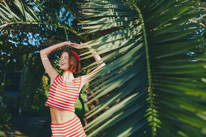 A cheerful woman in a striped crop top enjoys radiant sunlight among lush palm fronds, dancing with carefree energy and a bright, joyful expression in a vibrant outdoor scene.