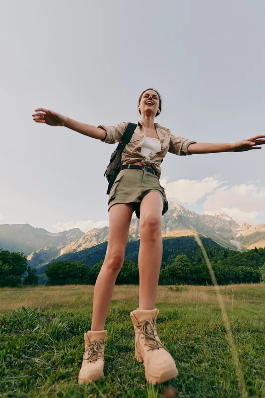 Young woman hiking outdoors with arms outstretched enjoying nature, wearing casual clothes and boots, mountain landscape background, bright daylight, active lifestyle