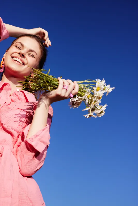 Young woman with a joyful expression holding wildflowers, wearing a stylish pink outfit against a clear blue sky, symbolizing spring and happiness