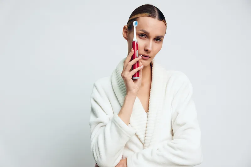 Young woman wearing a white bathrobe holding an electric toothbrush near her face on a light background, concept of dental care, hygiene, and morning routine with soft lighting.