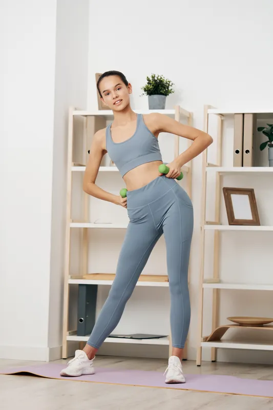 Woman in athletic gray outfit performs a side hip swing with light dumbbells in a bright home studio, showcasing fitness routine, balance, and confident movement beside minimalist shelves.