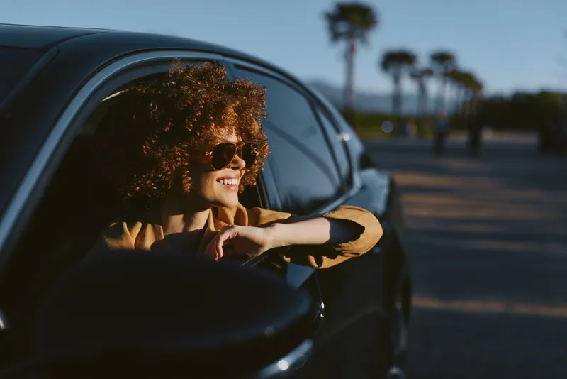 Woman with curly hair leans from car window, wearing sunglasses, enjoying a sunny drive, retro film color, palm trees in the background.