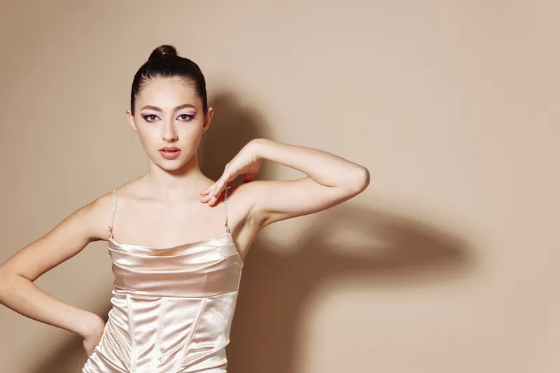 Elegant woman with sleek bun poses wearing a shiny beige satin dress. Minimal makeup highlights her sharp features. Studio portrait on a soft beige background.