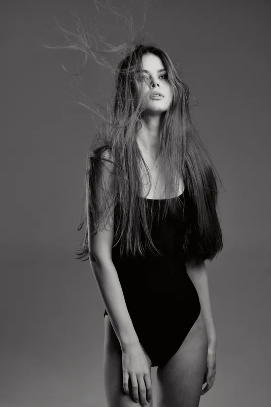 Black and white woman with long hair posing confidently in studio with a neutral gray background and soft lighting.