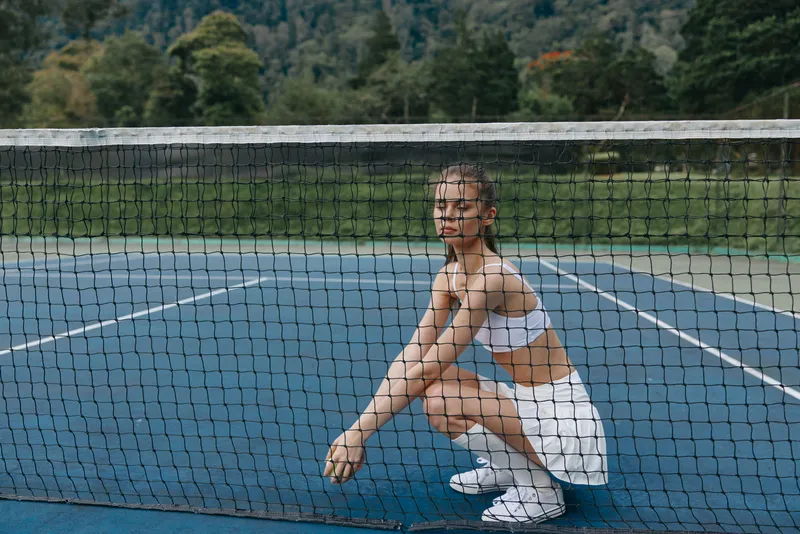 Athletic woman in sporty attire squatting near tennis net, displaying focus and determination on sunny outdoor court