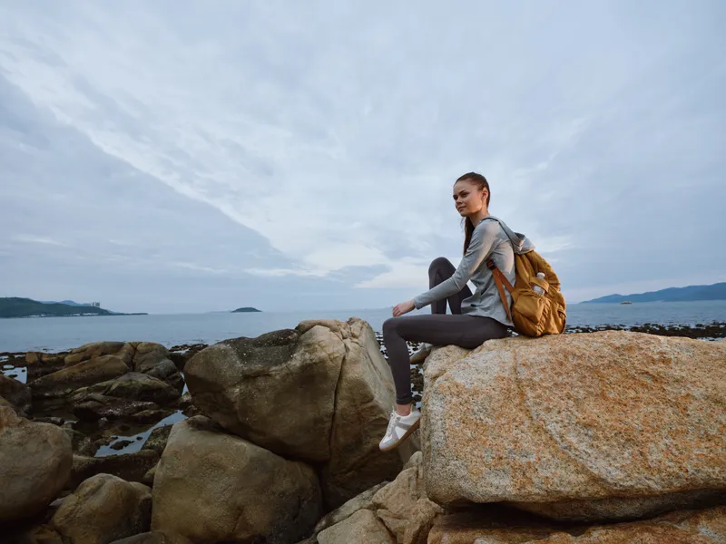 A young woman with a backpack sitting on rocks by the sea, showcasing adventure, exploration, and outdoor lifestyle in a serene coastal setting