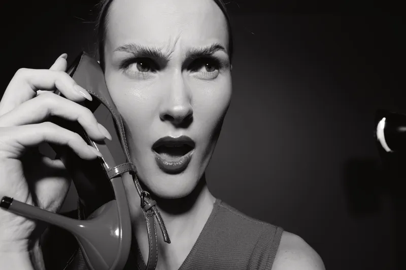 Young white woman with light skin holds high heel shoe near face with confused expression in black and white photo.