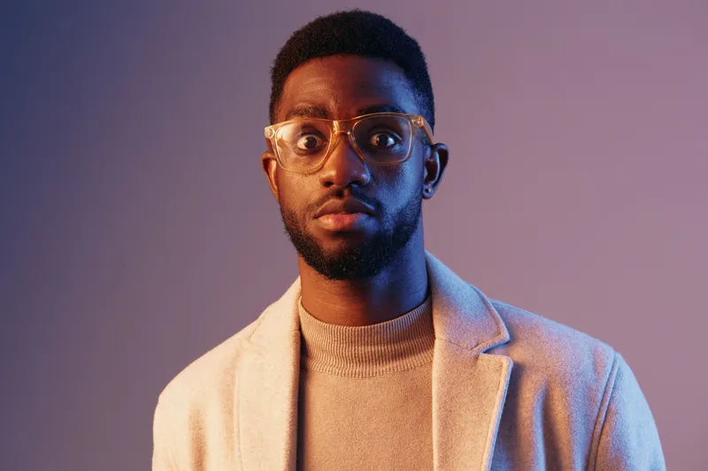 Stylish young African man in glasses, wearing a light coat and a sweater, expressing curiosity against a gradient background, perfect for modern fashion concepts