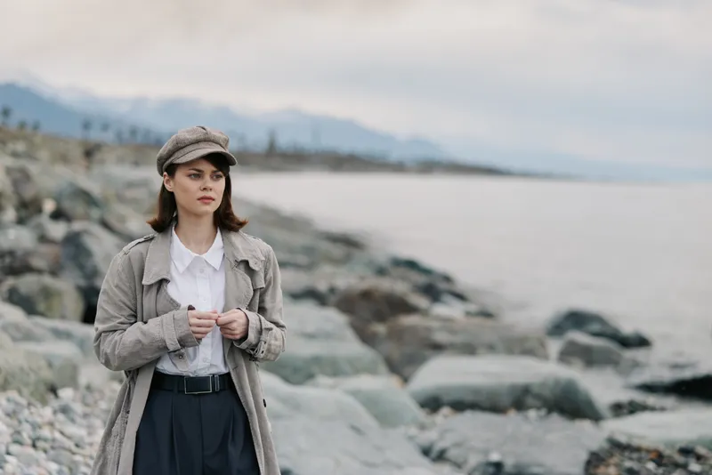 Young woman standing on rocky shore wearing knit cardigan and cap, looking into the distance. Overcast sky and calm sea in background. Concept of autumn fashion and contemplation.