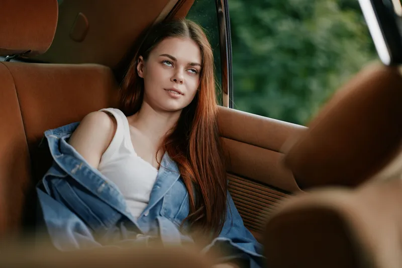 A young woman with long brown hair relaxing in a car, wearing a white tank top and blue denim jacket, showcasing a serene expression against a lush green background.
