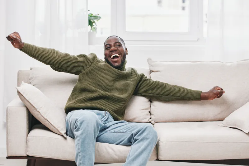 Happy African American man enjoying leisure time on a cozy couch, wearing a green sweater and jeans in a bright, airy living room