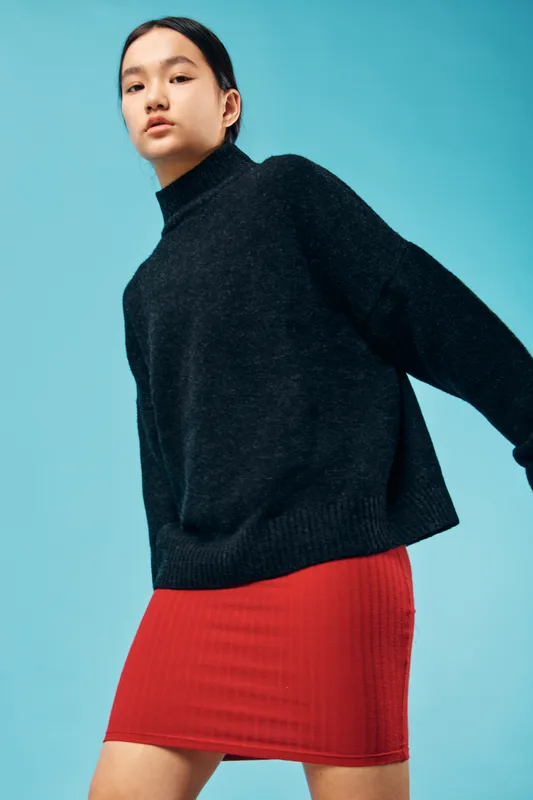 Young woman posing in studio wearing black oversized sweater and red skirt, minimalistic fashion look with clean blue background, casual style and confident expression.