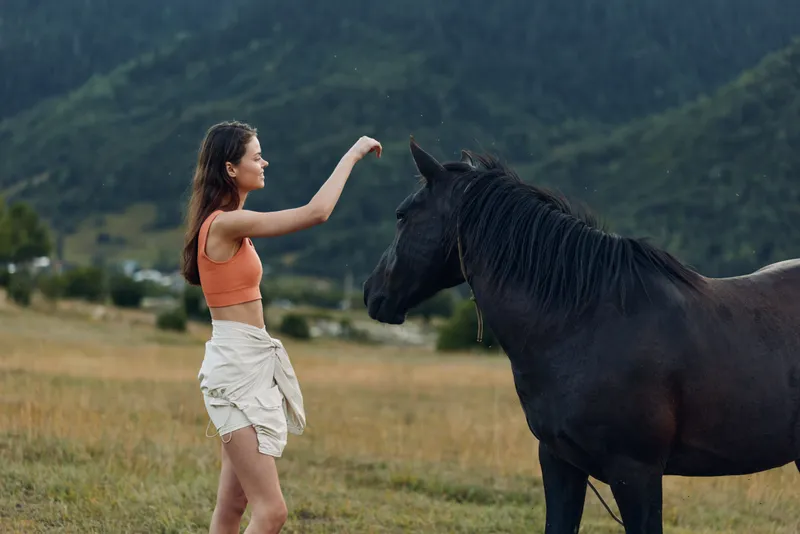 A young woman in an orange sleeveless top and light shorts stands in a wide open field, reaching toward a calm horse as the breeze moves the grass, creating a peaceful outdoor moment.