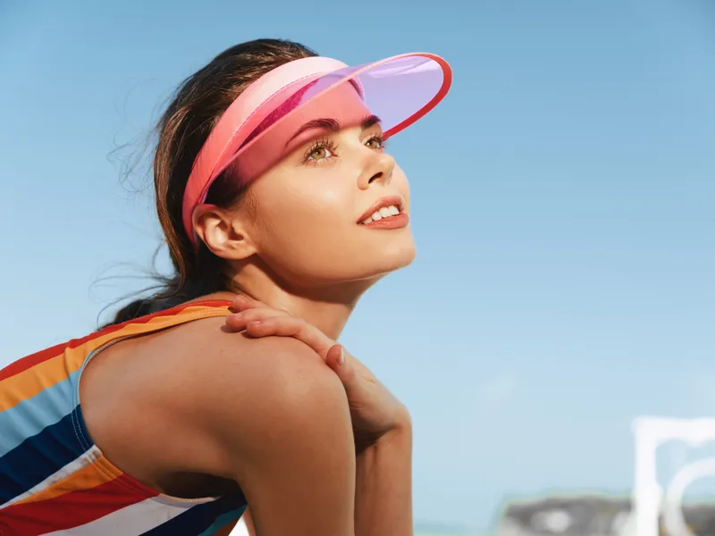 Smiling woman in a colorful striped swimsuit and pink visor gazes toward the sun, arms folded on her shoulders, framed by a clear blue sky and seaside backdrop for a bright summer moment.